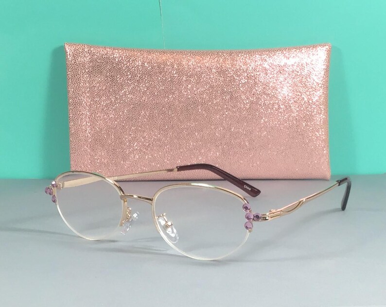 May include: Gold-rimmed spectacles with oval lenses and purple accents. The glasses are set against a rose gold glitter clutch. The frames have a delicate design, and the lenses are clear.