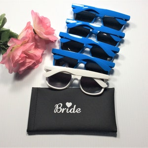Puede incluir: Un conjunto de gafas de sol azules y blancas, con lentes oscuras, junto con un estuche negro. El estuche lleva la palabra "Bride" y un s&iacute;mbolo de coraz&oacute;n. Tambi&eacute;n hay rosas rosas.