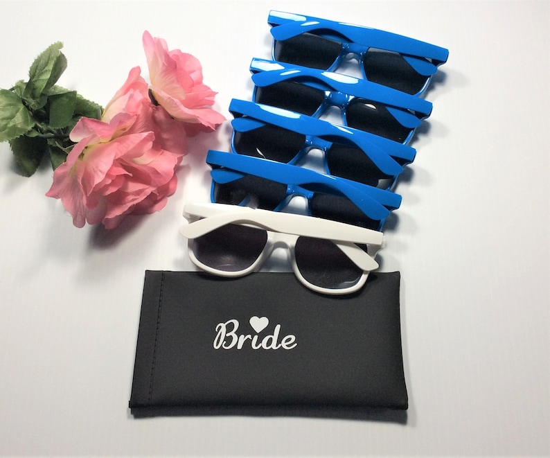 Puede incluir: Un conjunto de gafas de sol azules y blancas, con lentes oscuras, junto con un estuche negro. El estuche lleva la palabra "Bride" y un s&iacute;mbolo de coraz&oacute;n. Tambi&eacute;n hay rosas rosas.