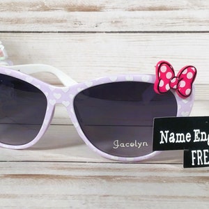 May include: Purple cat-eye sunglasses with white heart print and a pink bow with white polka dots. The sunglasses have a white frame and gray lenses. The text "Jacelyn" is written on the right lens. A black label with the text "Name Engraved FREE" is in the lower right corner of the image.