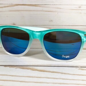 Puede incluir: Gafas de sol de color verde azulado y blanco con lentes espejados azules. Las gafas tienen la palabra "Suga" impresa en la lente derecha.