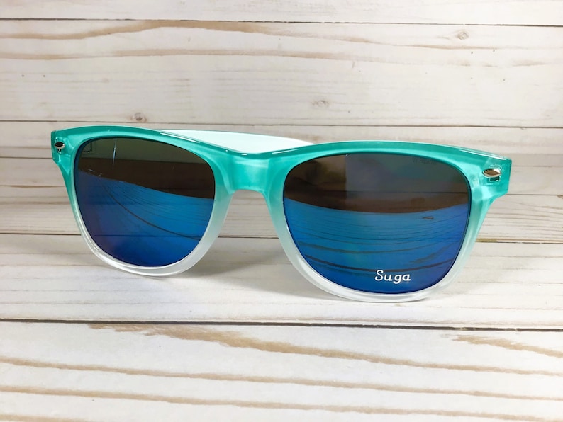 Puede incluir: Gafas de sol de color verde azulado y blanco con lentes espejados azules. Las gafas tienen la palabra "Suga" impresa en la lente derecha.