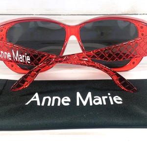 May include: Red sunglasses with dark lenses and the name "Anne Marie" printed on the temples. The sunglasses are paired with a black carrying case, also displaying the name "Anne Marie".