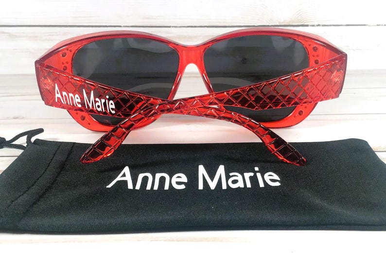 May include: Red sunglasses with dark lenses and the name "Anne Marie" printed on the temples. The sunglasses are paired with a black carrying case, also displaying the name "Anne Marie".