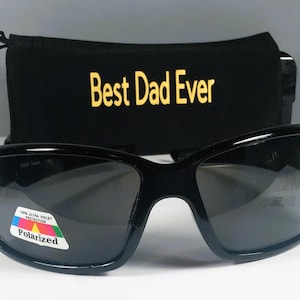Puede incluir: Gafas de sol negras con lentes oscuras y una pegatina polarizada con un gr&aacute;fico colorido y el texto 100% ULTRA VIOLET PROTECTION. Detr&aacute;s de las gafas hay una bolsa de tela negra con el texto amarillo Best Dad Ever impreso en ella.