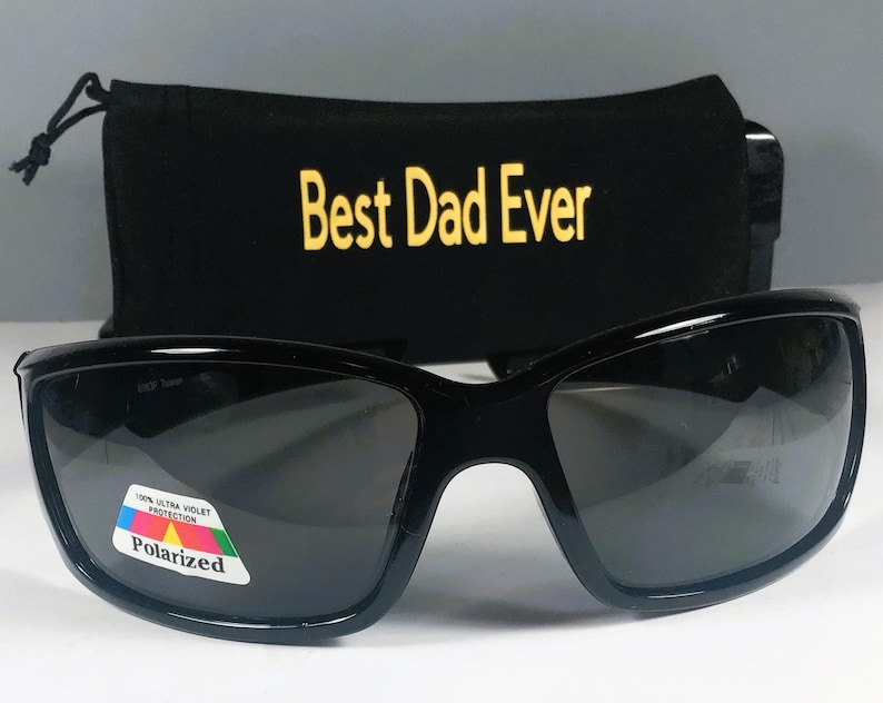 Puede incluir: Gafas de sol negras con lentes oscuras y una pegatina polarizada con un gr&aacute;fico colorido y el texto 100% ULTRA VIOLET PROTECTION. Detr&aacute;s de las gafas hay una bolsa de tela negra con el texto amarillo Best Dad Ever impreso en ella.