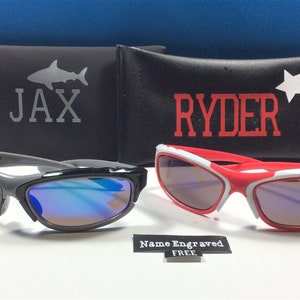 Peut inclure: Deux paires de lunettes de soleil avec des &eacute;tuis personnalis&eacute;s. Les lunettes de soleil sont noires et grises avec des verres bleus et rouges et blanches avec des verres violets. Les &eacute;tuis sont noirs avec un requin et le nom "JAX" sur l'un et une &eacute;toile et le nom "RYDER" sur l'autre. Le texte "Nom grav&eacute; GRATUIT" est &eacute;galement visible.