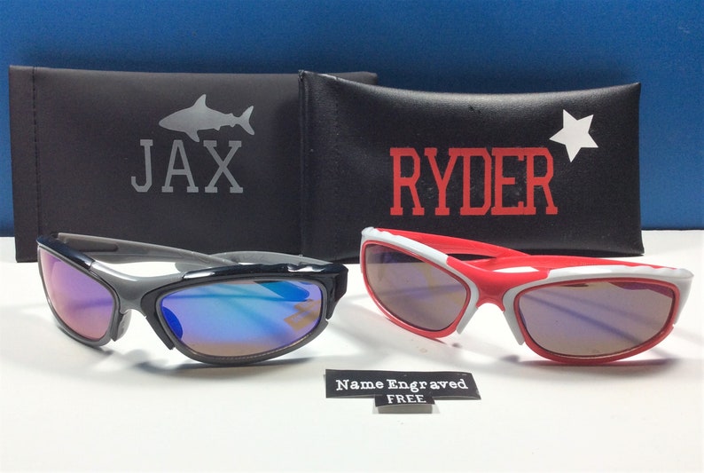 Peut inclure: Deux paires de lunettes de soleil avec des &eacute;tuis personnalis&eacute;s. Les lunettes de soleil sont noires et grises avec des verres bleus et rouges et blanches avec des verres violets. Les &eacute;tuis sont noirs avec un requin et le nom "JAX" sur l'un et une &eacute;toile et le nom "RYDER" sur l'autre. Le texte "Nom grav&eacute; GRATUIT" est &eacute;galement visible.