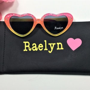 May include: Pink and yellow heart-shaped sunglasses with black lenses and rhinestones. The sunglasses are in a black case with the name "Raelyn" and a pink heart printed on it.