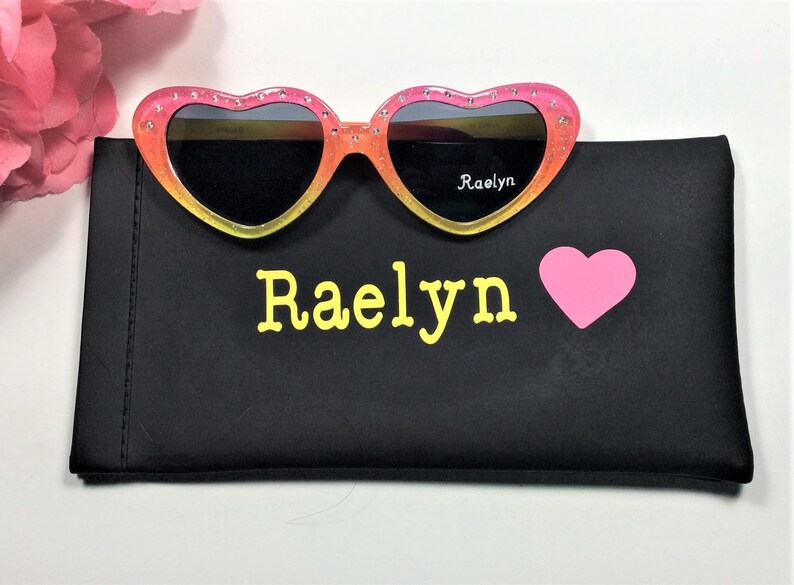 May include: Pink and yellow heart-shaped sunglasses with black lenses and rhinestones. The sunglasses are in a black case with the name "Raelyn" and a pink heart printed on it.