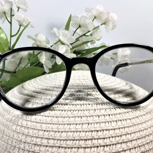 K&ouml;nnte beinhalten: Schwarz umrandete Brille mit klaren Gl&auml;sern und dekorativen, dunkel schimmernden Kristallverzierungen an den &auml;u&szlig;eren Ecken des Rahmens. Die B&uuml;gel sind d&uuml;nn und metallisch und verbinden sich an den verzierten Scharnieren mit dem Rahmen.