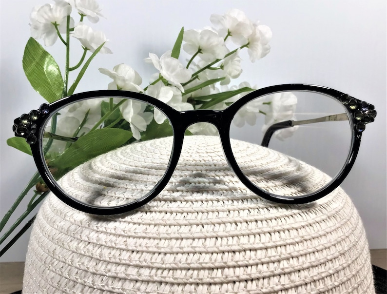 K&ouml;nnte beinhalten: Schwarz umrandete Brille mit klaren Gl&auml;sern und dekorativen, dunkel schimmernden Kristallverzierungen an den &auml;u&szlig;eren Ecken des Rahmens. Die B&uuml;gel sind d&uuml;nn und metallisch und verbinden sich an den verzierten Scharnieren mit dem Rahmen.