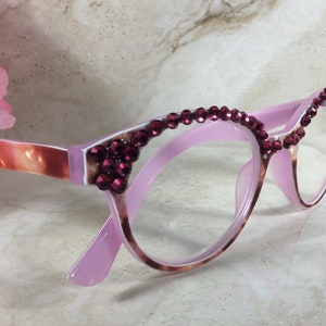 K&ouml;nnte beinhalten: Rosafarbene Brille im Cat-Eye-Stil, verziert mit weinroten Strasssteinen entlang der oberen R&auml;nder. Die B&uuml;gel weisen ein marmoriertes Muster in Rosa- und Braunt&ouml;nen auf, was einen stilvollen Look verleiht. Ein modisches Accessoire.