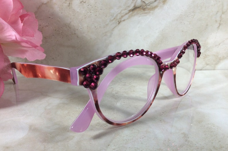 K&ouml;nnte beinhalten: Rosafarbene Brille im Cat-Eye-Stil, verziert mit weinroten Strasssteinen entlang der oberen R&auml;nder. Die B&uuml;gel weisen ein marmoriertes Muster in Rosa- und Braunt&ouml;nen auf, was einen stilvollen Look verleiht. Ein modisches Accessoire.