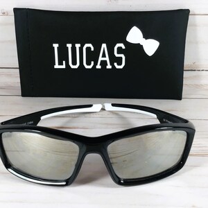 May include: Black sunglasses with mirrored lenses and white accents. The sunglasses come with a black carrying case that has the name "LUCAS" and a white bow tie printed on it.
