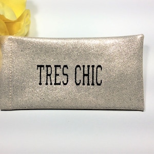 Pu&ograve; includere: Una pochette rettangolare dall'aspetto metallico con un motivo maculato. Il testo "TRES CHIC" &egrave; stampato in nero sul davanti. Un fiore giallo &egrave; visibile nell'angolo in alto a sinistra.