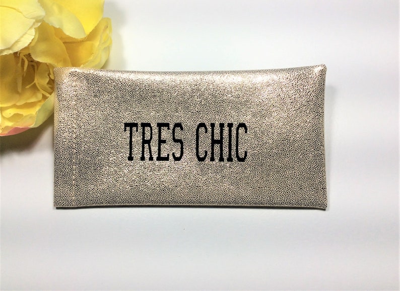 Pu&ograve; includere: Una pochette rettangolare dall'aspetto metallico con un motivo maculato. Il testo "TRES CHIC" &egrave; stampato in nero sul davanti. Un fiore giallo &egrave; visibile nell'angolo in alto a sinistra.
