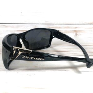 May include: Black sunglasses with a sleek, modern design. The sunglasses feature dark lenses and a glossy black frame with silver accents and the brand name "XLOOP". The sunglasses are resting on a light-colored wooden surface.