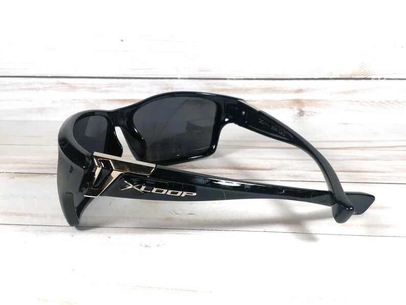 May include: Black sunglasses with a sleek, modern design. The sunglasses feature dark lenses and a glossy black frame with silver accents and the brand name "XLOOP". The sunglasses are resting on a light-colored wooden surface.