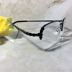 May include: Black rimless spectacles with clear lenses. The temples are black with decorative black accents. The glasses are resting on a light-coloured surface, with a yellow flower in the background.