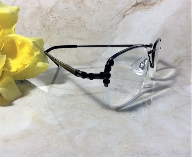 May include: Black rimless spectacles with clear lenses. The temples are black with decorative black accents. The glasses are resting on a light-coloured surface, with a yellow flower in the background.