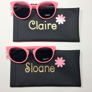 May include: Two pink sunglasses with black lenses in black cases. The cases have a pink flower and the name "Claire" and "Sloane" written in gold lettering.