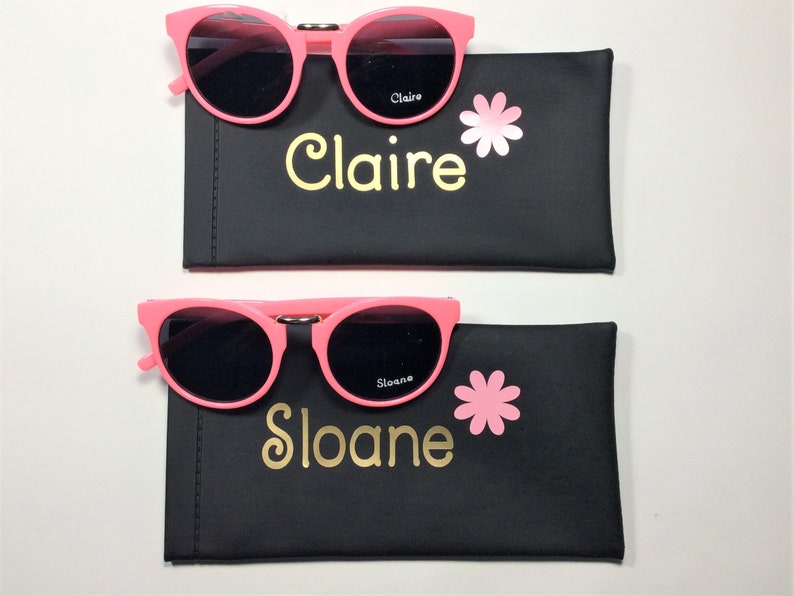May include: Two pink sunglasses with black lenses in black cases. The cases have a pink flower and the name "Claire" and "Sloane" written in gold lettering.