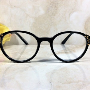 May include: Black round-frame eyeglasses with clear lenses. The frames have gold-colored stud accents on the outer corners. The glasses are set against a light-colored, marble-like background.