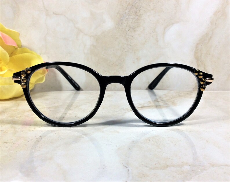 May include: Black round-frame eyeglasses with clear lenses. The frames have gold-colored stud accents on the outer corners. The glasses are set against a light-colored, marble-like background.