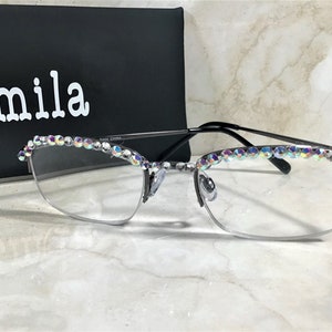 May include: Half-rim eyeglasses with a black frame and clear lenses. The top of the frame is embellished with iridescent rhinestones. The glasses are in front of a black box with the name "Camila" in white.