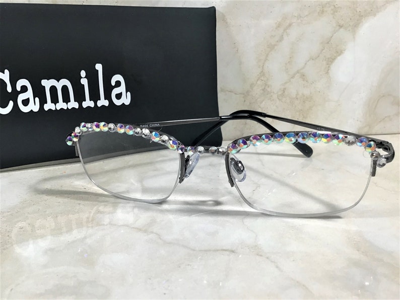 May include: Half-rim eyeglasses with a black frame and clear lenses. The top of the frame is embellished with iridescent rhinestones. The glasses are in front of a black box with the name "Camila" in white.