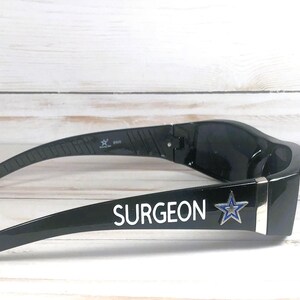 May include: Black sunglasses with the word "SURGEON" in white lettering and a blue star logo. The sunglasses have a sleek, modern design and are set against a light wood-grain background.
