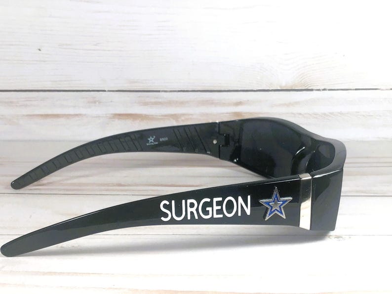May include: Black sunglasses with the word "SURGEON" in white lettering and a blue star logo. The sunglasses have a sleek, modern design and are set against a light wood-grain background.