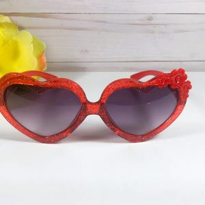 May include: Red heart-shaped sunglasses with glitter and a red flower embellishment.