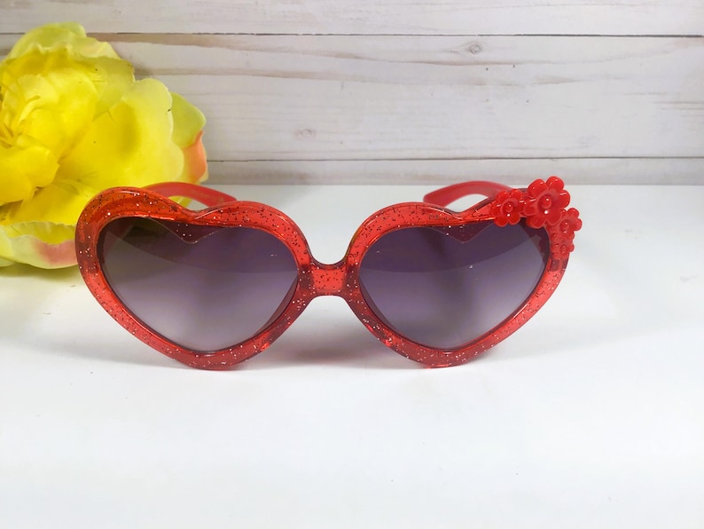 May include: Red heart-shaped sunglasses with glitter and a red flower embellishment.