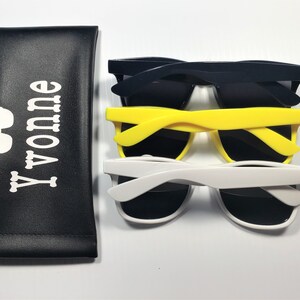 May include: A black rectangular pouch with the white text Yvonne and a small glasses icon, accompanied by three pairs of sunglasses in dark blue, bright yellow, and white, each featuring dark lenses and thick plastic frames.