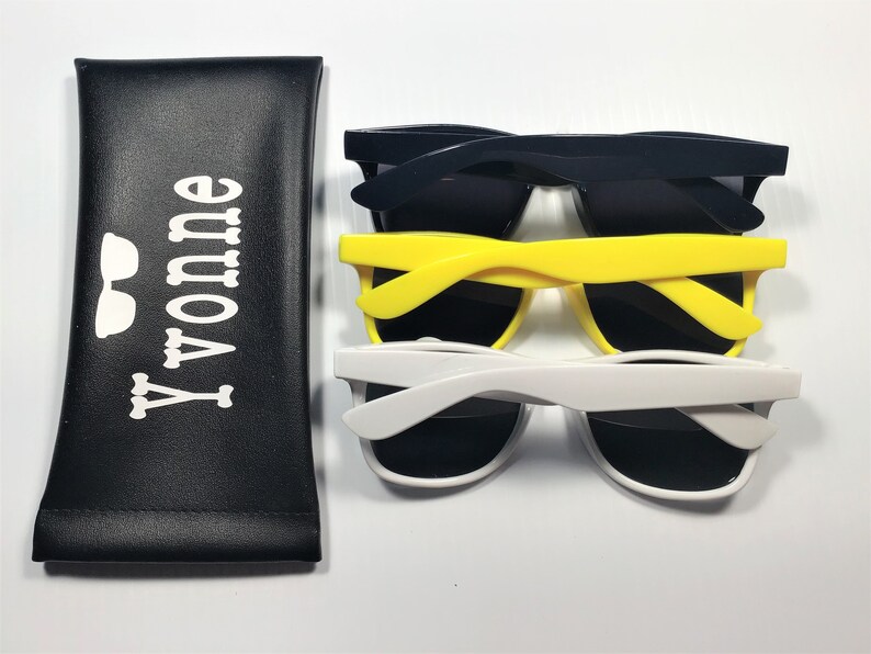 May include: A black rectangular pouch with the white text Yvonne and a small glasses icon, accompanied by three pairs of sunglasses in dark blue, bright yellow, and white, each featuring dark lenses and thick plastic frames.