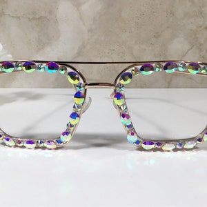 Peut inclure: Une paire de lunettes de forme carr&eacute;e avec une monture en or rose et des strass iris&eacute;s autour des verres.