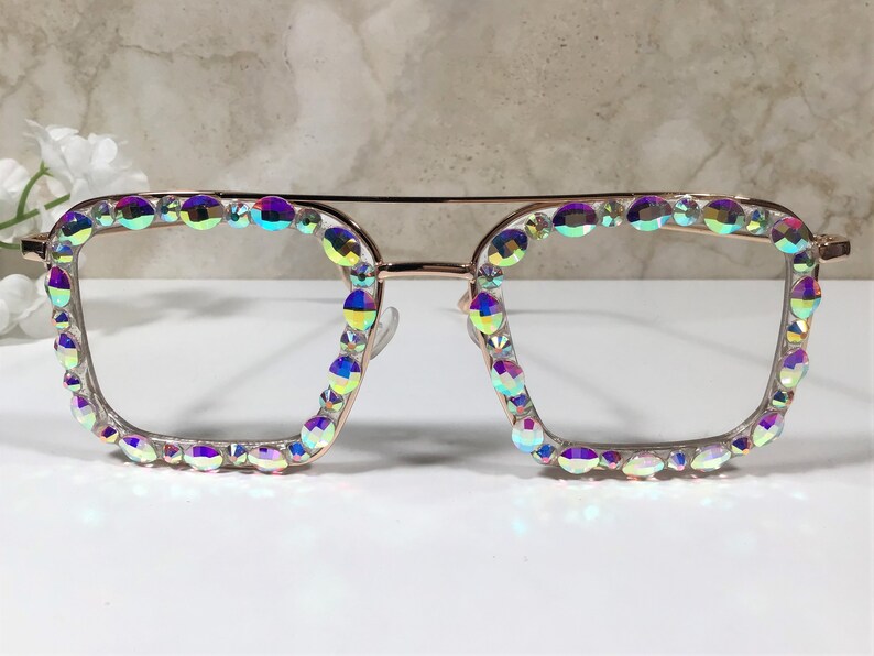 Peut inclure: Une paire de lunettes de forme carr&eacute;e avec une monture en or rose et des strass iris&eacute;s autour des verres.