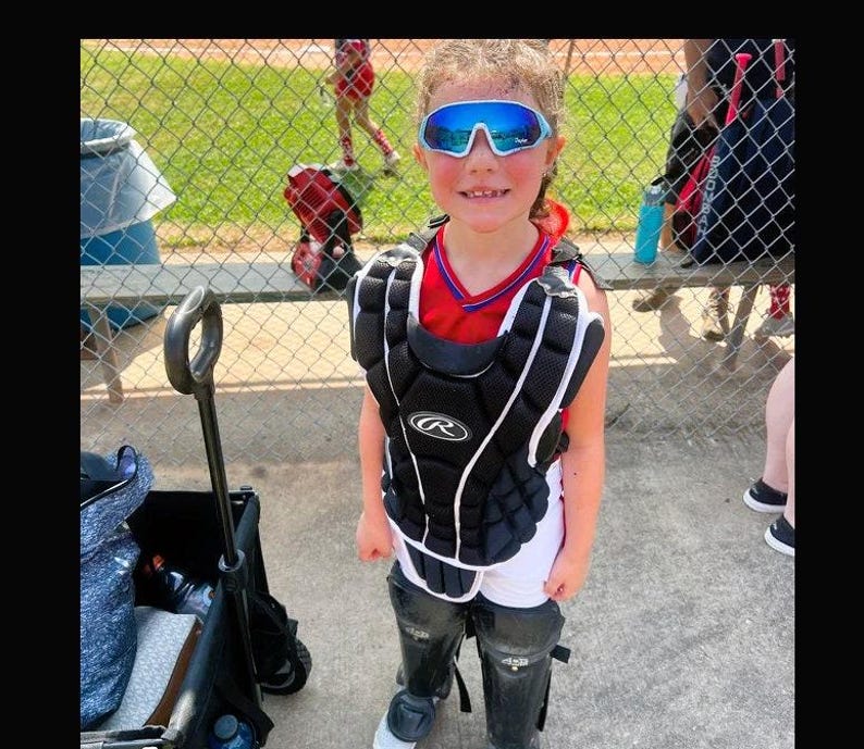 May include: A youth baseball catcher wearing a black Rawlings chest protector over a red and blue jersey, white shorts, and black leg guards. The catcher wears blue-tinted mirrored sports sunglasses with a white frame.