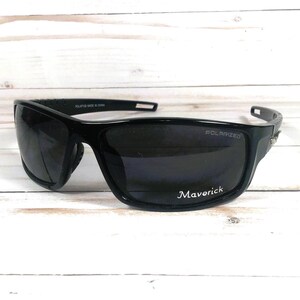 May include: Black polarized sunglasses with the word "Maverick" printed on the lens. The sunglasses have a sporty design with a thick frame and a curved shape. The lenses are dark, and the frame is glossy.