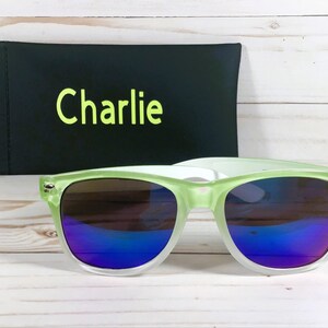 Puede incluir: Gafas de sol verdes y blancas con lentes espejados azules. Las gafas de sol vienen con una funda negra que dice "Charlie" en ella.