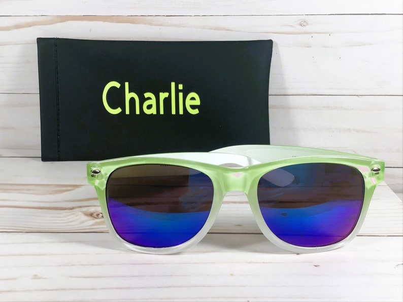 Puede incluir: Gafas de sol verdes y blancas con lentes espejados azules. Las gafas de sol vienen con una funda negra que dice "Charlie" en ella.
