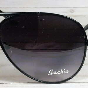 Pu&ograve; includere: Occhiali da sole neri a goccia con lenti sfumate. Gli occhiali hanno una sottile montatura nera e il nome "Jackie" &egrave; stampato in bianco sulla lente.