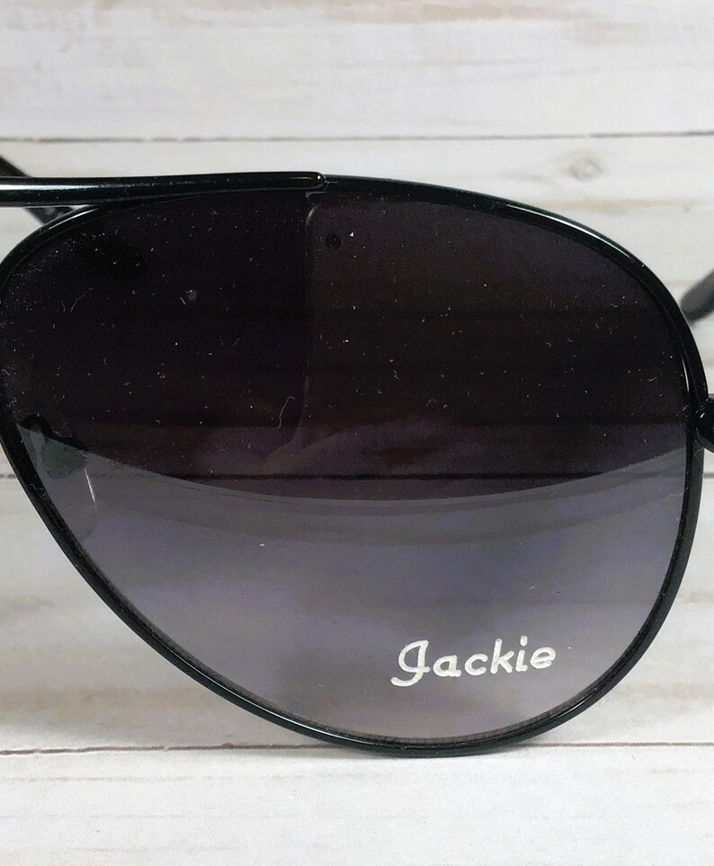 Pu&ograve; includere: Occhiali da sole neri a goccia con lenti sfumate. Gli occhiali hanno una sottile montatura nera e il nome "Jackie" &egrave; stampato in bianco sulla lente.