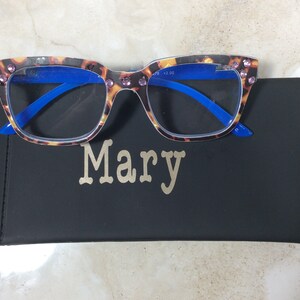 May include: Reading glasses with a tortoiseshell pattern frame and blue arms, adorned with pink rhinestones. The glasses rest on a black case with the name "Mary" in gold lettering.