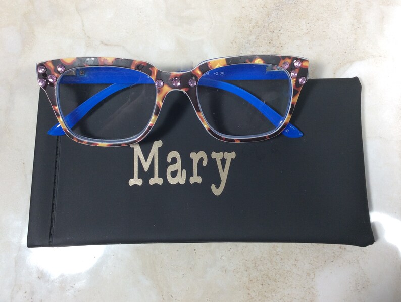 May include: Reading glasses with a tortoiseshell pattern frame and blue arms, adorned with pink rhinestones. The glasses rest on a black case with the name "Mary" in gold lettering.