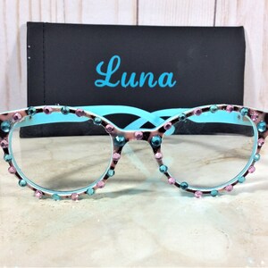 May include: Cat-eye style eyeglasses with a light blue frame and embellished with pink and teal rhinestones. The lenses are clear. The frame has a brown and white pattern. A black case with the name "Luna" in teal script is behind the glasses.