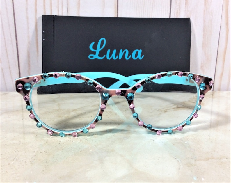 May include: Cat-eye style eyeglasses with a light blue frame and embellished with pink and teal rhinestones. The lenses are clear. The frame has a brown and white pattern. A black case with the name "Luna" in teal script is behind the glasses.