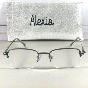 May include: Rimless eyeglasses with a silver frame and decorative black accents at the temples. The glasses are in front of a white, textured eyeglass case with the name "Alexia" in black script.